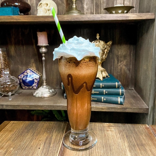Um copo transparente decorado com chocolate, com uma bebida no estilo milkshake de cor marrom coberta por chantilly e com um canudo de papel listrado em branco e verde.