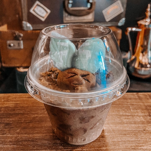 Chocolate cremoso com bolacha, parecendo terra e uma trufa de chocolate com marcas que parecem olhos e boca e folhas de chocolate branco saindo pela parte de cima da trufa. Tudo em um pote transparente com tampa arredondada.