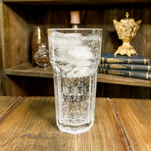 Um copo transparente de água com gás e gelo. Atrás, uma estante com vários objetos coloridos.