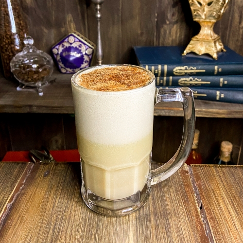 Uma caneca transparente com várias camadas em tons de caramelo, com canela e noz moscada por cima. Ao fundo, uma estante com vários itens coloridos.