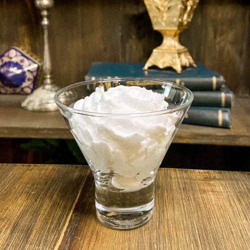 Uma taça transparente com uma porção de chantilly. Ao fundo, uma prateleira com diversos itens coloridos.