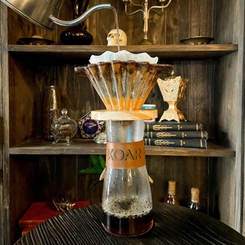 Um porta-filtro ondulado com filtro de papel sobre um decanter de vidro cheio até a metade de café, na parte mais fina no decanter há um sleeve de couro