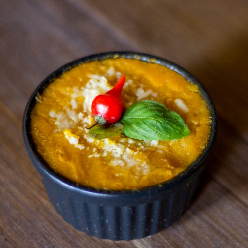 Um ramequim preto com creme de abóbora; por cima queijo ralado e decoração de pimenta dedo de moça.