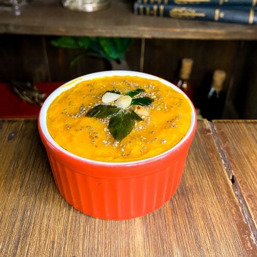Um ramequim vermelho com creme de abóbora; por cima queijo ralado e decoração de manjericão.