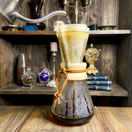 Uma jarra transparente com café já filtrado. O filtro cônico de camada tripla pode ser visto na parte de cima da jarra, que tem um anel central de madeira amarrado por uma cordinha de couro.
