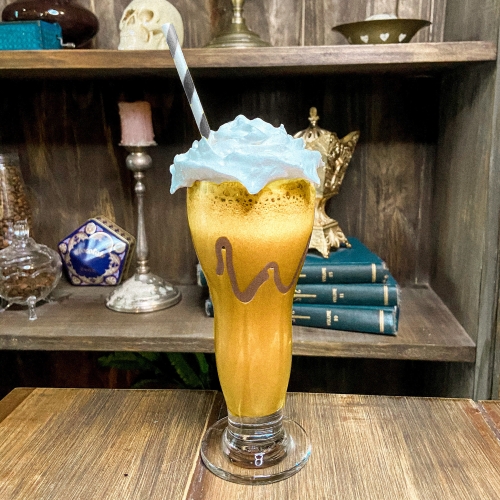 Um copo transparente decorado com chocolate, com uma bebida no estilo milkshake de cor laranja coberta por chantilly e com um canudo de papel listrado em branco e preto.