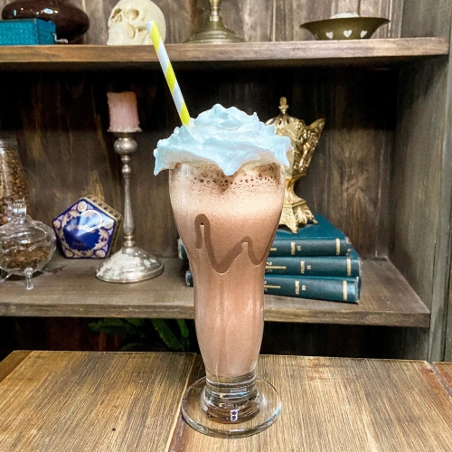 Um copo transparente decorado com chocolate, com uma bebida no estilo milkshake de cor marrom clara coberta por chantilly e com um canudo de papel listrado em branco e amarelo.