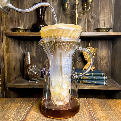 Uma jarra grande e transparente, com gelo na parte central e filtro com café na parte superior, café sendo passado com água quente por uma chaleira que vemos uma parte aparecendo ao lado.