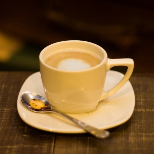 Uma xícara branca pequena com café com um pinguinho de leite, em cima de um pires com uma colher com uma balinha amarela com manchinhas marrom