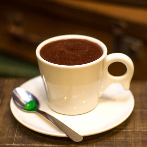Uma xícara branca com chocolate quente dentro, em um pires com uma colher com uma balinha verde 
