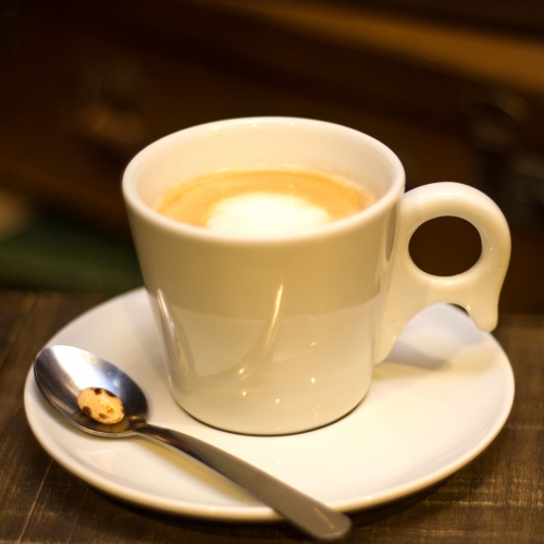Uma xícara branca com café e um pingo de leite, em cima de um pires com uma colher com uma balinha alaranjada