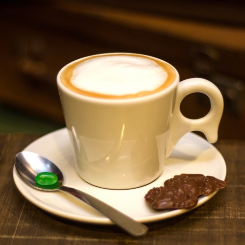 Uma xícara branca com café com leite dentro, em cima de um pires com um sapinho de chocolate