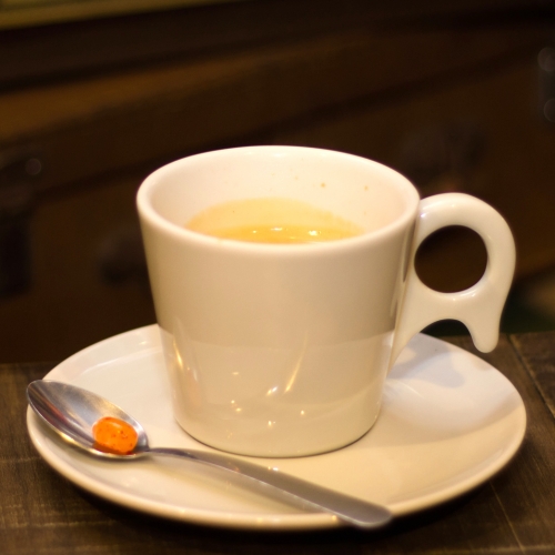 Uma xícara branca com café dentro, em cima de um pires com uma colher com uma balinha laranja