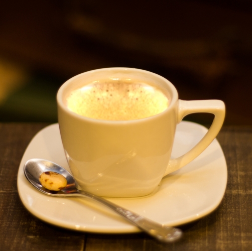 Uma xícara branca pequena com café dentro, em cima de um pires com uma colher com uma balinha alaranjada