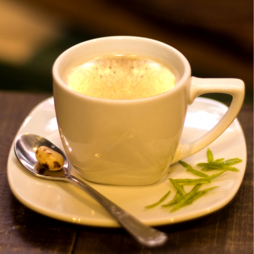 Uma xícara branca pequena com café dentro, em cima de um pires com raspas de limão e uma colher com uma balinha
