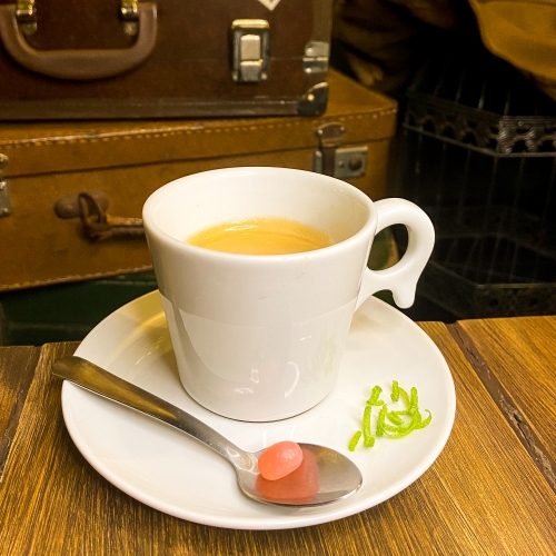 Uma xícara branca com café dentro, em cima de um pires com raspas de limão e uma colher com uma balinha