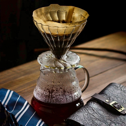 Uma jarra transparente com café já filtrado; sobre ela há um porta-filtro cônico também transparente. Dentro dele vemos um filtro com café. Na mesa, ainda há um cardápio, uma gravata e uma varinha.