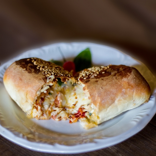 Pão baguete coberto por gergelim, cortado ao meio, com queijo, tomate seco e rúcula de recheio, sobre um prato branco